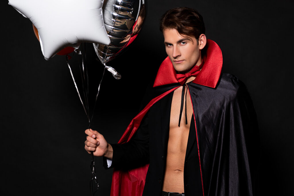 A person in a Dracula costume holding star-shaped balloons stands proudly against a black background, embracing their unique identity and celebrating the vibrant spirit of the LGBTQIA+ community.