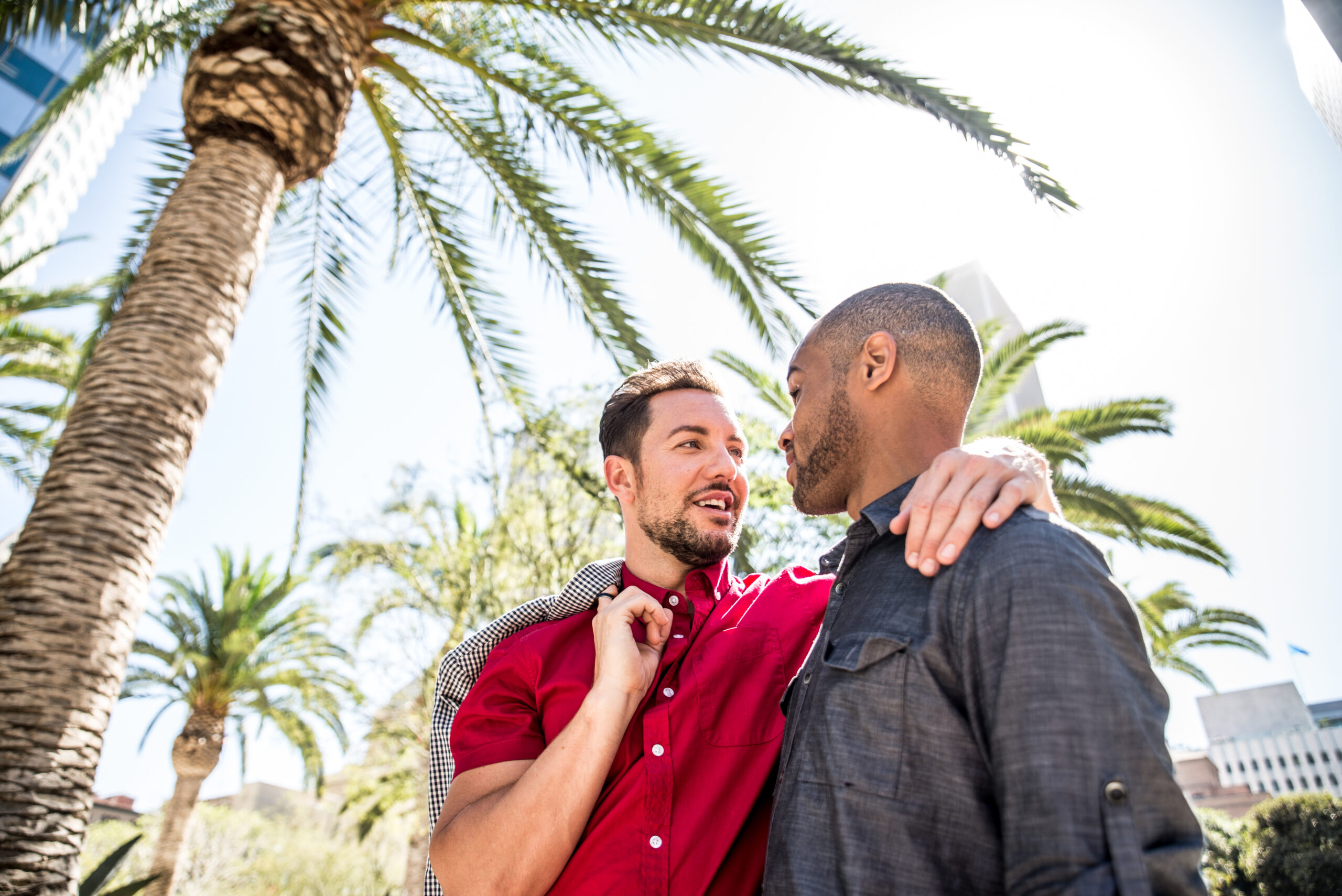 Two people smiling at each other under palm trees on a sunny day, their bond evident as one person has an arm around the other, embodying the warmth of relationships.