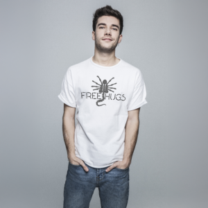 A man wearing a white "Free Hugs" T-shirt, featuring a scorpion design, stands with his hands in his pockets against a gray backdrop, exuding a touch of sci-fi charm.