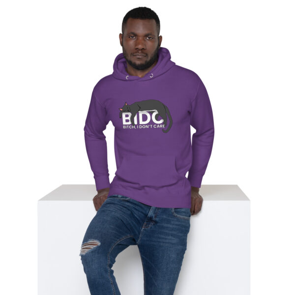 Man wearing a BIDC Hoodie, featuring a cat design and text, leaning against a white surface.