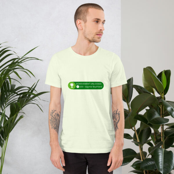 A person stands near green plants, wearing a light-colored t-shirt featuring the green graphic text "Achievement: Gaymer Boyfriend.