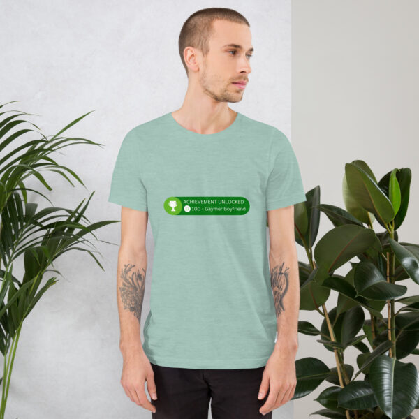 A person wearing a light green T-shirt featuring the text "Achievement: Gaymer Boyfriend" stands near green plants.