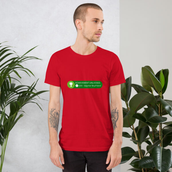 A man in a red "Achievement: Gaymer Boyfriend" t-shirt, standing next to potted plants.