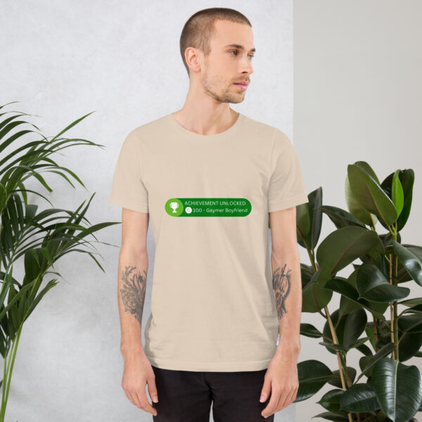 A person wearing the "Achievement: Gaymer Boyfriend" beige t-shirt, featuring a green text design, stands near indoor plants.