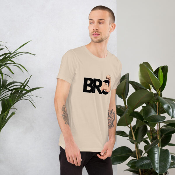 A man stands near green plants, wearing a beige Bro t-shirt featuring a fist graphic.