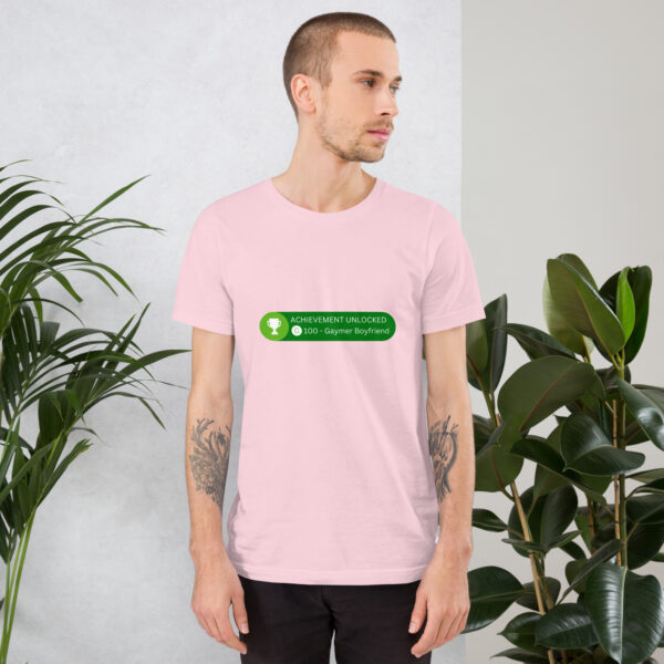 A person in a light pink T-shirt with the "Achievement: Gaymer Boyfriend" design, featuring green text that reads "Unlocked 0:00 - Gaymer Boyfriend," stands near potted plants.