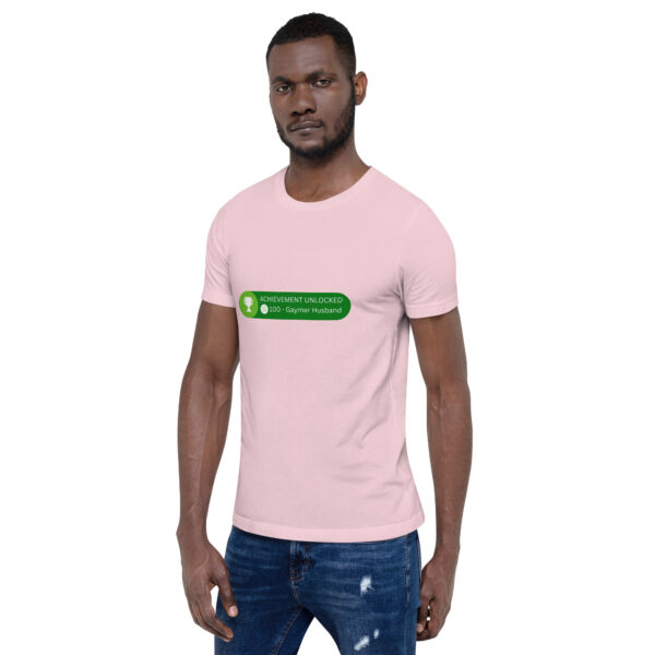 A man wearing a pink t-shirt with the text "ACHIEVEMENT UNLOCKED. 100G - Gaymer Husband" printed on the front, standing against a plain background.