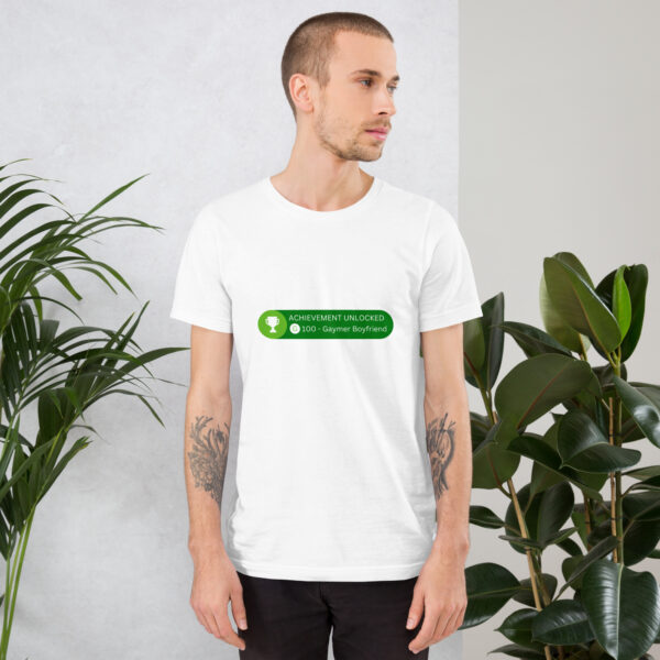 A person wearing a white T-shirt with the design "Achievement: Gaymer Boyfriend" is standing near tall green plants.