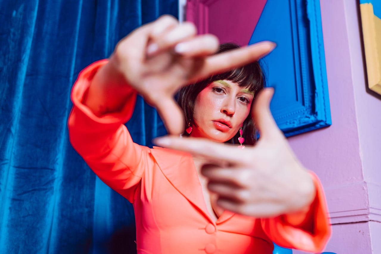 Person in a bright pink outfit frames their face with their hands against a colorful background.