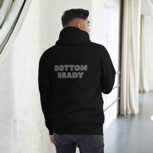 A person wearing a Bottom Ready Hoodie, with "BOTTOM READY" printed on the back, stands indoors by a white brick wall and curtains.