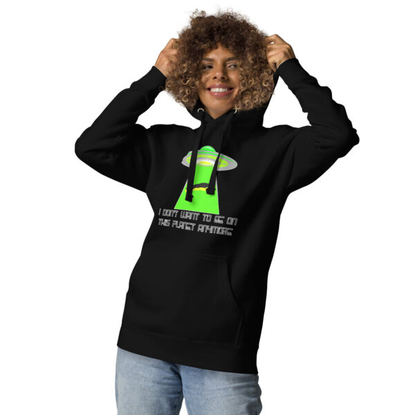 A person smiling and holding their curly hair is wearing the "I don't want to be on this planet anymore" hoodie, featuring a black color with a green alien spaceship design and text.