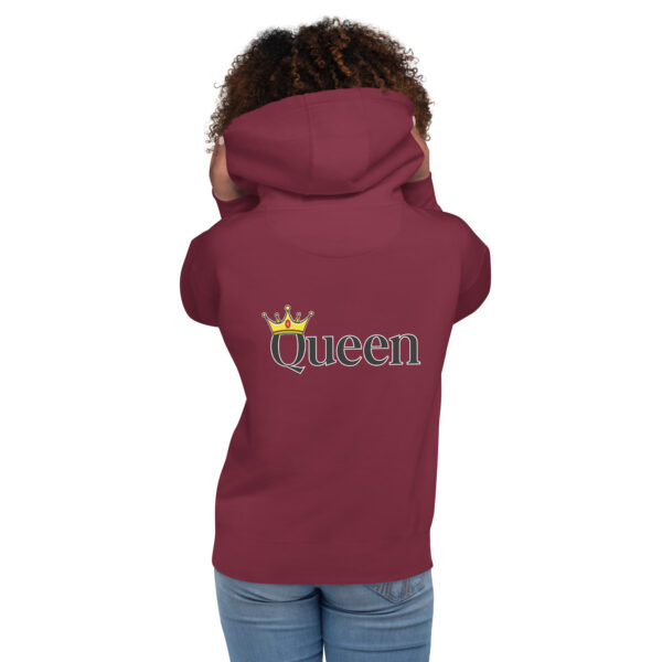 A person is wearing the Queen Hoodie, which features a maroon color with the word "Queen" and a crown graphic on the back, while standing with both hands on their hood.