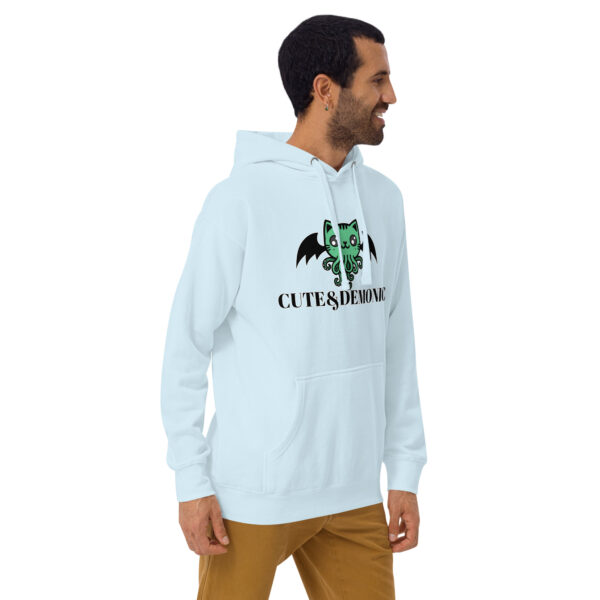 A man wearing the Cute and Demonic Hoodie in light blue, featuring "CUTE & DEMONIC" and a cartoon green creature design, is standing and smiling.