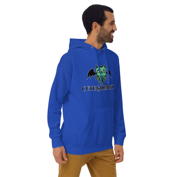 A person smiles and looks to the side, wearing the "Cute and Demonic Hoodie," which features a blue design with a cartoon cat graphic and the text "CUTE & DEMONIC," paired with brown pants.