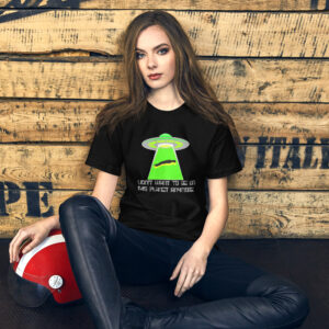 A woman sitting on the floor against a wooden wall is wearing a black shirt featuring the "I Don't Want To Live On This Planet Anymore" design, which includes a green alien and text. Beside her on the floor sits a red and white helmet, evoking the theme of cosmic adventure.