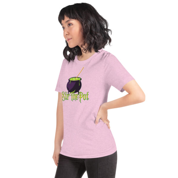 A person in a light pink "Stir The Pot" shirt featuring a cauldron graphic stands in a side pose with hands on hips.