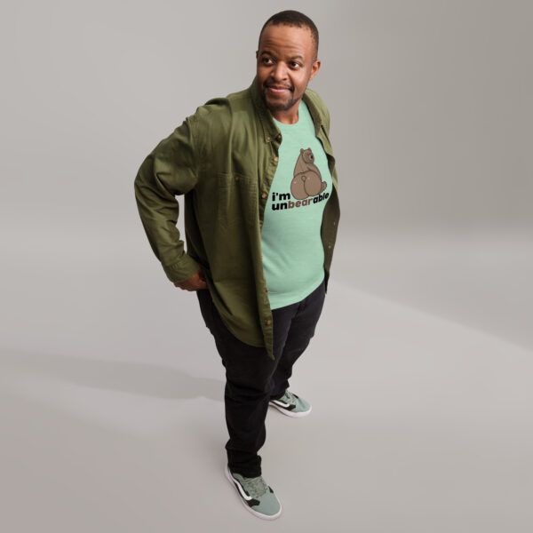 A man in a green "I'm unbearable" shirt, paired with a jacket, black pants, and sneakers, stands on a light gray floor looking to his right.