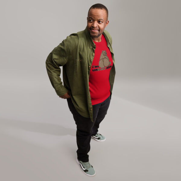 A man stands with a slight smile, wearing a green jacket over an "I'm unbearable" red graphic t-shirt, dark pants, and sneakers.