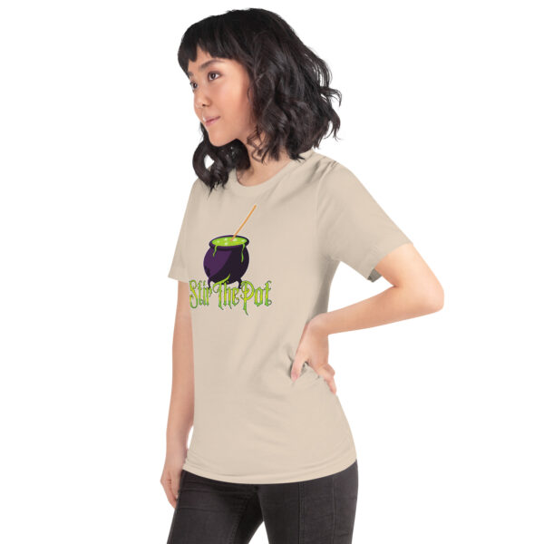 A person is wearing the "Stir The Pot" beige T-shirt featuring a graphic of a purple cauldron with text.